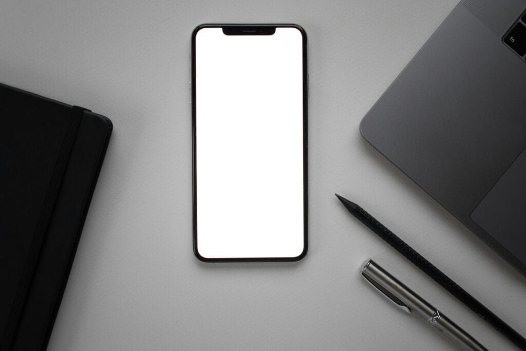 blank cellphone screen, pen, and laptop on a desk